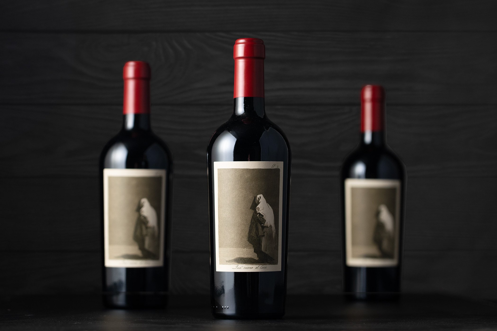 Three wine bottles with black bodies and red caps, displayed against a black wooden background, with vintage-style labels featuring mountain illustrations.