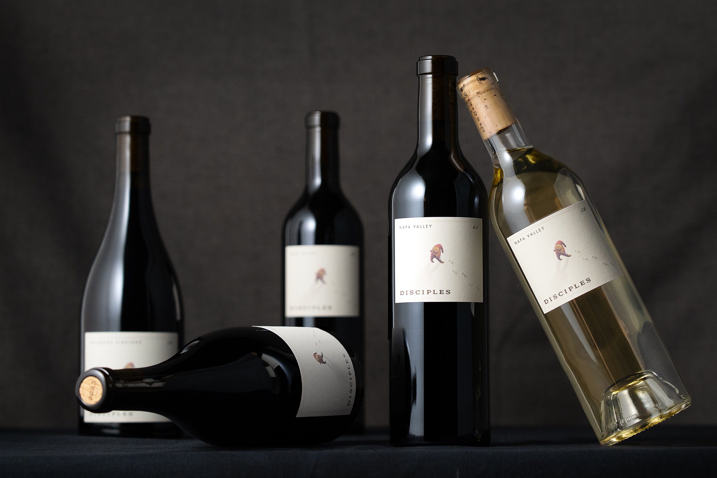 Several bottles of wine, some upright and some lying down, on a dark surface against a dark background. The labels on the bottles indicate they are from Napa Valley and have the name 'Disciples' with a small bear illustration.